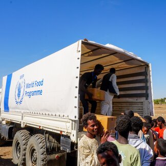 World Food Programme truck with staff distributing aid boxes to people gathered outside; WFP logo visible on the truck's side.