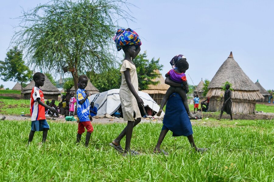 A South Sudanese woman carrying a baby and her three other children stride through the grass, with a thatched hut in the backdrop. Photo: WFP/Michael Tewelde 