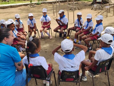  Children wearing white caps and blue scarves sit in a circle outdoors with WFP staff, participating in an interactive group activity.