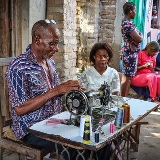 Person operating a sewing machine at an outdoor table with colourful spools of thread, while others sit and stand nearby against a brick wall.