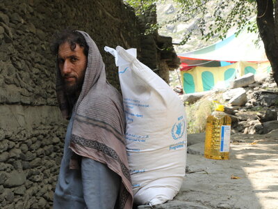 A person stands beside a large WFP food sack on a rocky path, with a bottle of cooking oil nearby and colourful tents visible in the background.