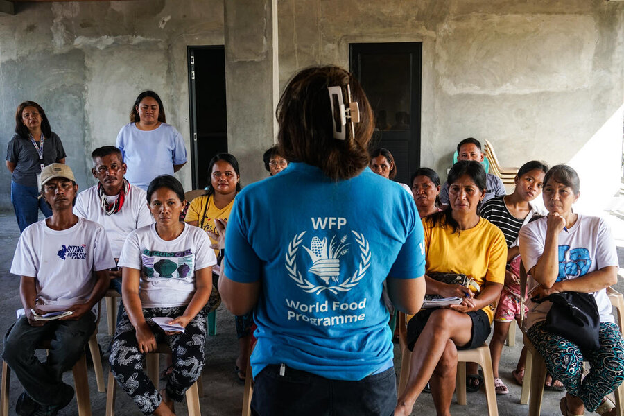 Around 12 people sitting in rows on chairs listen to a woman wearing a WFP-logo shirt with her back to the camera