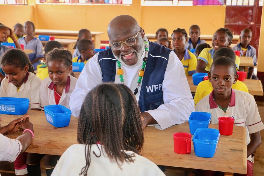 A man wearing a blue WFP vest and African beads sits in a primary school classroom in eastern Cameroon chatting with young students. Photo: WFP/Jordan Onana