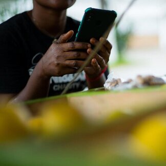 Person holding a smartphone at a market stall with yellow fruits and packaged goods in the foreground.