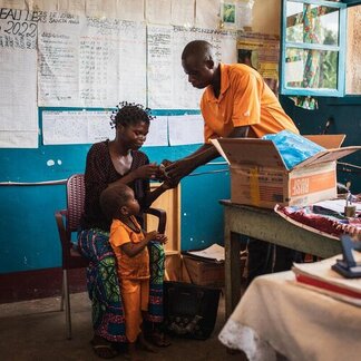 Health worker hands medicine to a seated adult with a child nearby in a clinic room with charts on the wall and supplies on a desk.