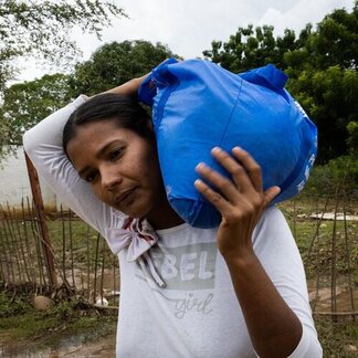 Person carries a large blue bag on their shoulder while standing outdoors near a rustic fence, trees and a parked car in the background.