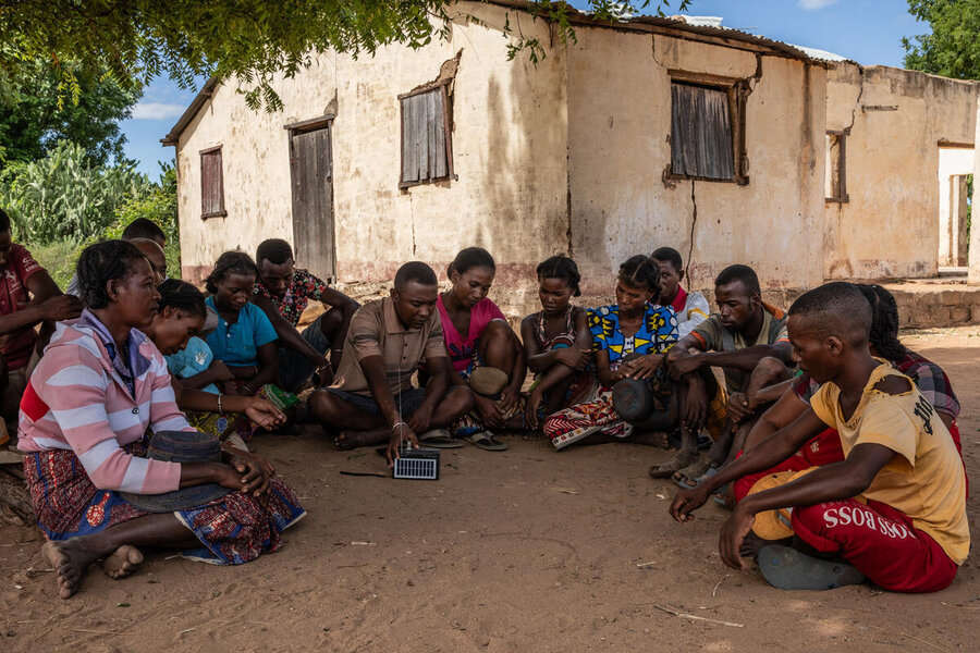 A large group of people sat on the ground in front of a village structure around a radio