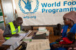 A man in a yellow vest writes in a logbook while a woman and her child watch. In the background, a WFP banner is visibl