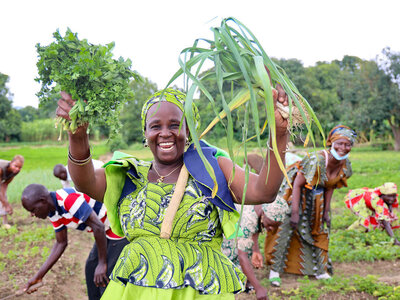 A smiling woman dressed in green is proudly holding  parsley and leek grown in the garden, with fellow farmers harvesting in the background.