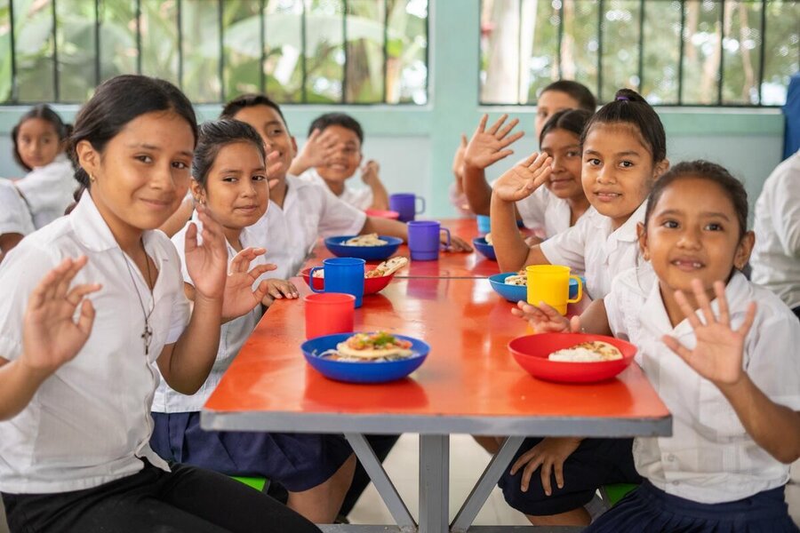 In Honduras, children at red a table at lunchtime at school wave to the camera