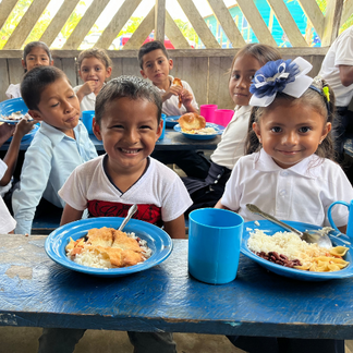 Children sitting at a wooden table in a school setting, eating plates of rice, beans, pasta, and a portion of fried food, with brightly coloured cups and bowls around them.